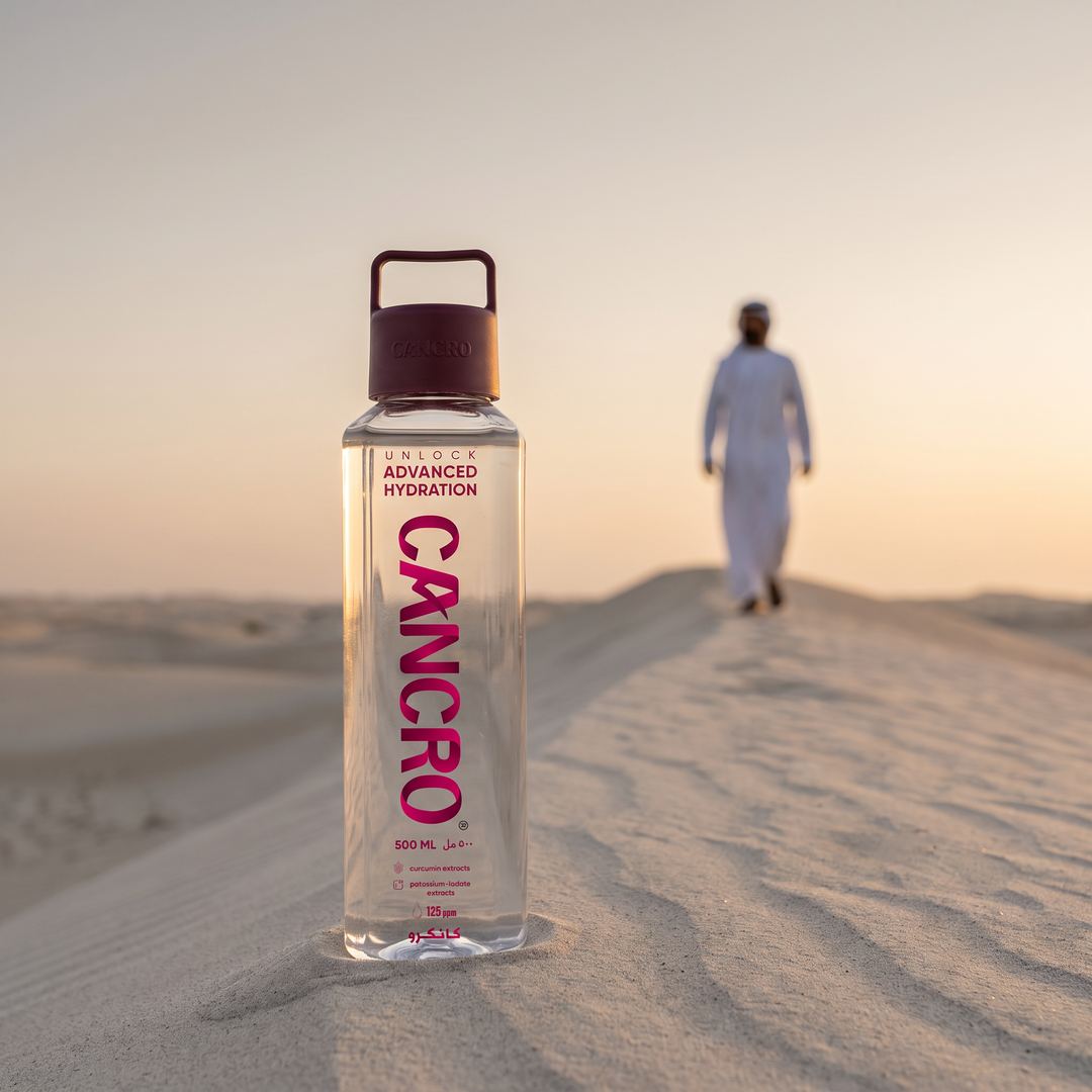A Cancro bottle standing in desert sand while a man in a kandura walks over the dune behind it