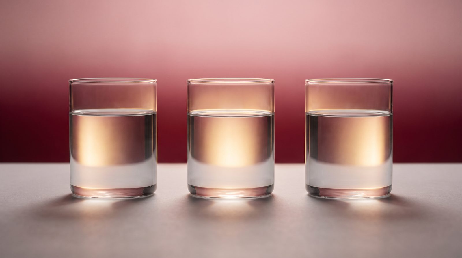 Three clear glass vessels of water lit from behind against a deep wine-red gradient